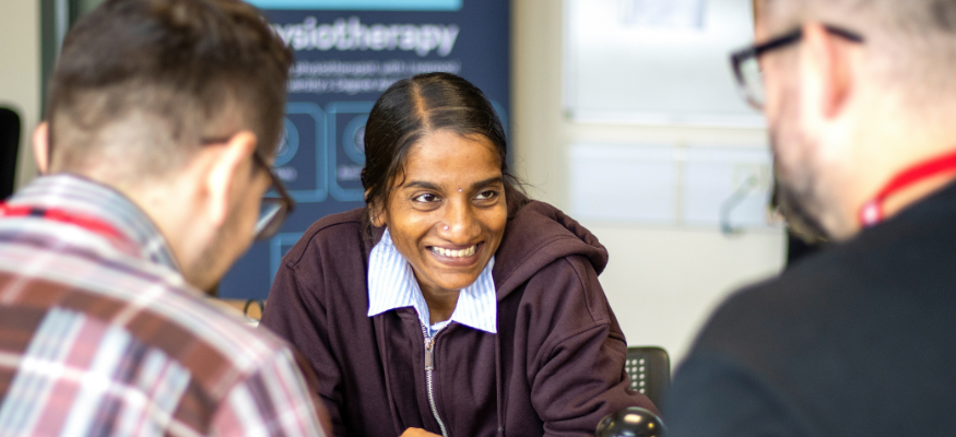A lecturer is sat at a desk, facing the camera and smiling at colleagues who are facing her, as they engage in conversation.